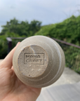 Hand holding a ceramic cup with 'Maha Craft' branding against a blurred natural background