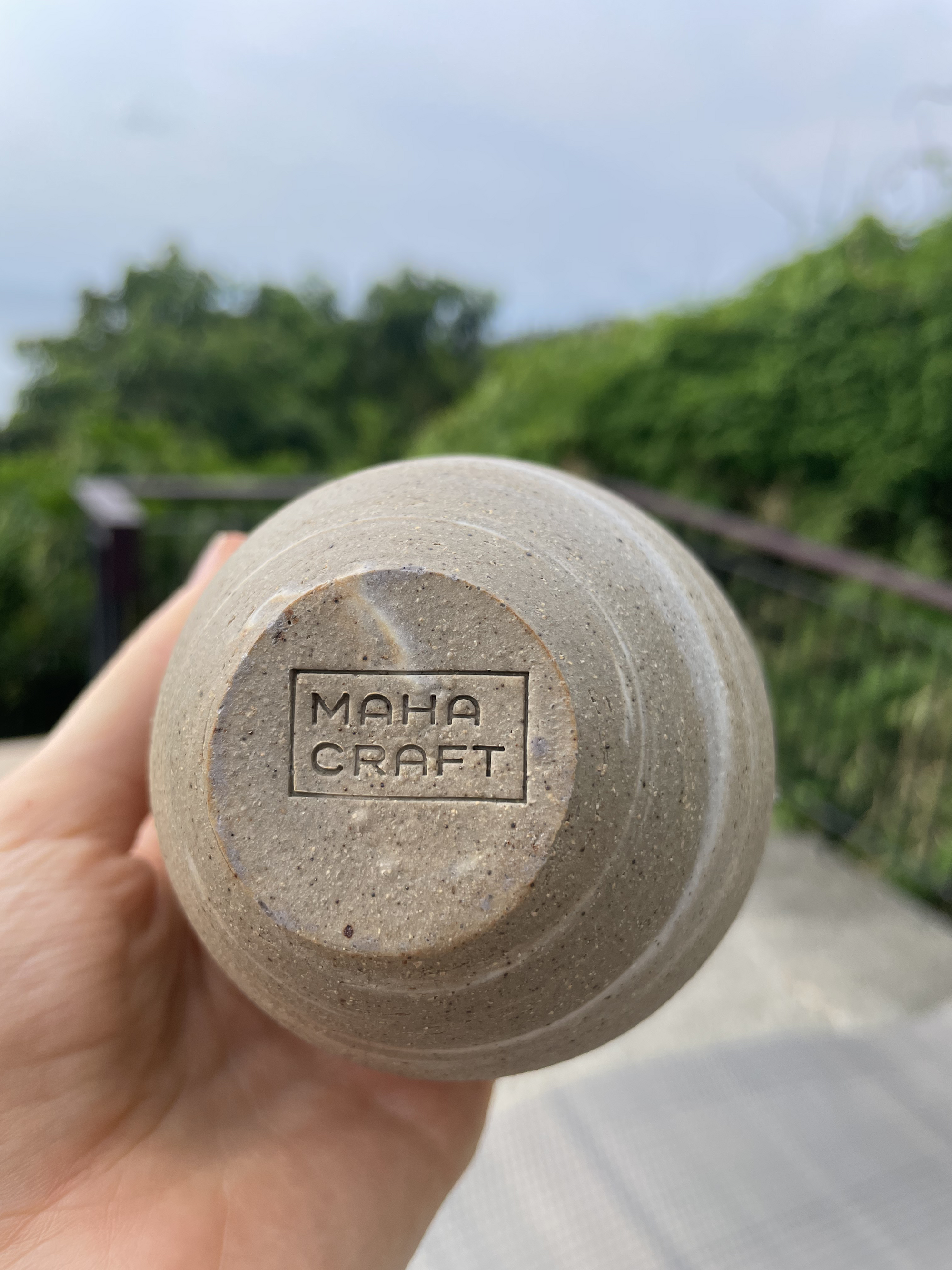 Hand holding a ceramic cup with 'Maha Craft' branding against a blurred natural background